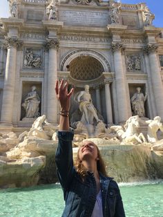 a woman standing in front of a fountain reaching up to the sky with her hands