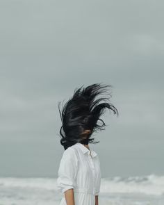 a woman standing on the beach with her hair blowing in the wind and looking at the ocean