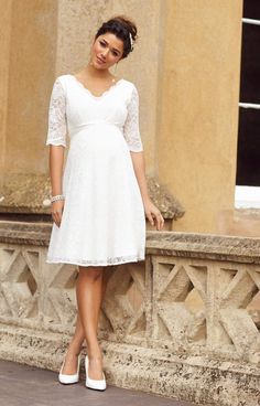 a woman in a white dress leaning against a wall with her hand on her hip