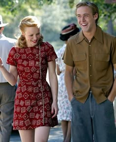 a woman in a red dress walking next to a man wearing a brown shirt and hat