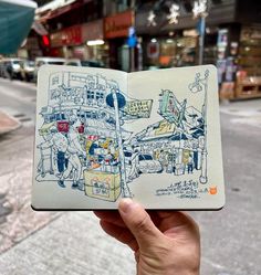 a hand holding up an open notebook with drawings on the pages in front of a city street