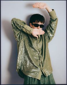 a man with sunglasses covering his face while standing in front of a white wall and holding his hands behind his head