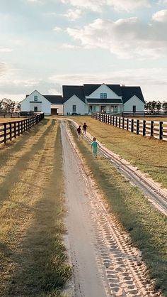 two people walking down a dirt road in front of a white house