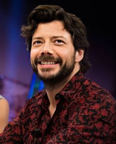 a man with a beard smiles at the camera while wearing a red and black shirt