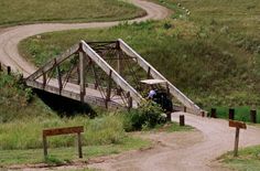 a small bridge over a dirt road in the middle of a grassy area with signs on either side