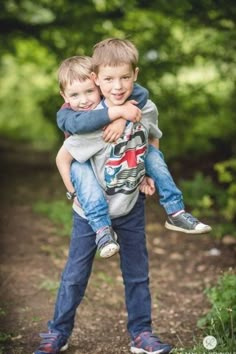 two young boys hugging each other on a dirt path in the middle of some trees