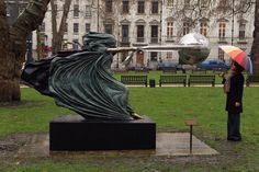 a woman holding an umbrella standing next to a statue in a park near a building