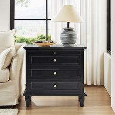 a night stand with a lamp on top of it next to a couch and window