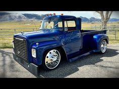 an old blue truck parked in a parking lot next to a tree and fence with mountains in the background