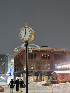 two people walking in the snow next to a clock on a pole with lights around it