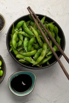 green beans and black sesame seeds in bowls with chopsticks next to them on a white surface