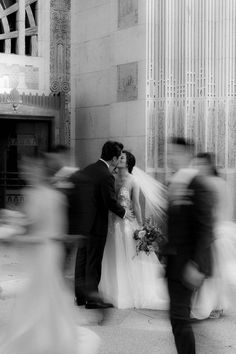 a bride and groom kissing in front of a building with many people walking by them