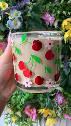 a hand holding a glass candle with cherries on it and flowers in the background