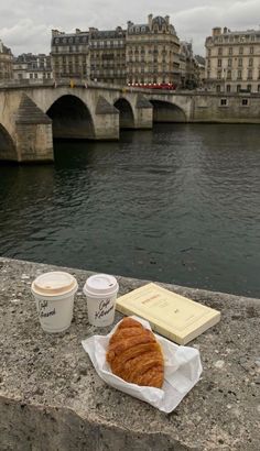 coffee and croissants are sitting on the edge of a bridge overlooking a body of water