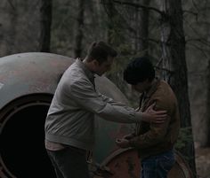 two men standing next to each other near a barrel in the woods with trees behind them