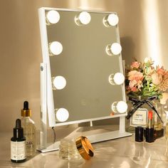 a vanity mirror with lights on it next to some bottles and flowers in front of the mirror