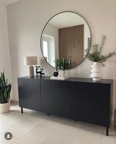 a large round mirror hangs on the wall above a black cabinet with flowers in it