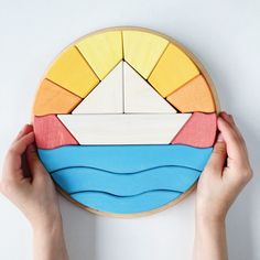 two hands are holding a colorful wooden sailboat on a white surface with blue, yellow, and red colors