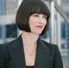 a woman with short hair wearing a black suit and gold necklace standing in front of stairs