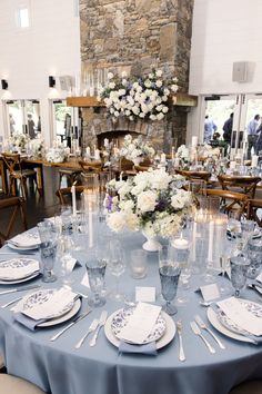 a table set up with plates, silverware and flowers in front of a stone fireplace