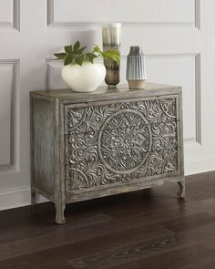 an ornate wooden cabinet with two vases on top and one plant in the corner