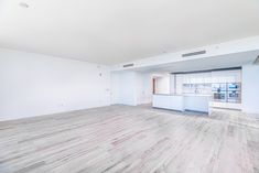 an empty living room with white walls and wood flooring in the foreground is a kitchen