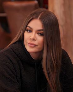 a woman with long brown hair wearing a black jacket and looking at the camera while sitting down