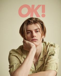 a young man sitting with his hand on his chin and looking at the camera while wearing a shirt that says ok