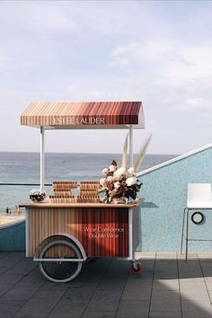 an ice cream cart on the beach with flowers in front of it and water behind it