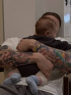 a man holding a baby with tattoos on his arms and shoulder, while sitting in a chair