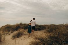 a man and woman are walking on the sand dunes together, with grass in the foreground