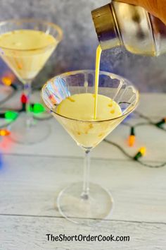 someone pouring something into a martini glass on a white table with christmas lights in the background