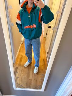 a young boy taking a selfie in the mirror with his cell phone while wearing an orange and green jacket