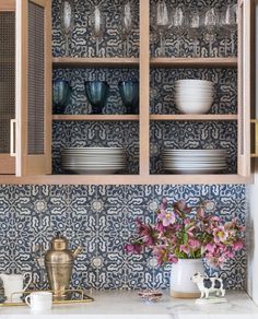 a cabinet with dishes and cups on it in a kitchen next to a vase filled with flowers