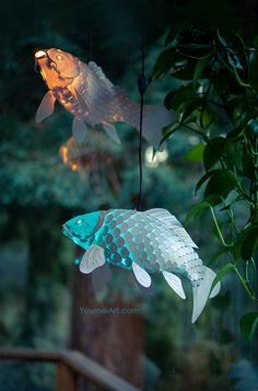 two fish hanging from strings in front of some trees and bushes with lights on them