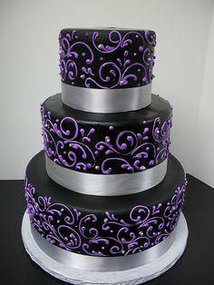 a three tiered cake with purple and silver decorations on the top, sitting on a black table