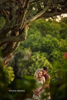 a man and woman standing under a tree in front of some green trees with their arms around each other