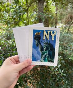 someone holding up a new york postcard with the statue of liberty on it in front of some trees