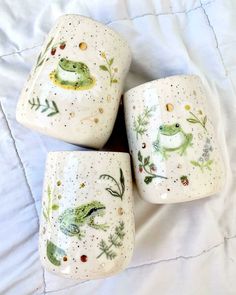 four ceramic cups with frog designs on them sitting on a bed together, one is green and the other is white