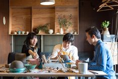 three people sitting at a table with cups and plates on it, looking at something in the distance