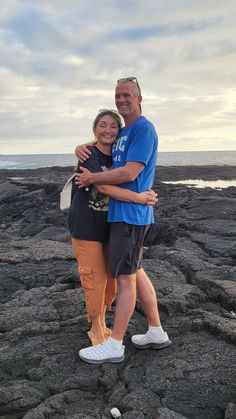 a man and woman standing on top of a rocky beach next to the ocean with their arms around each other