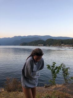 a woman standing next to a body of water with mountains in the backgroud