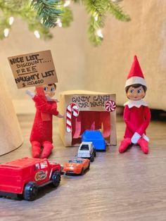 two elfs are sitting on the floor next to toy cars and a christmas tree