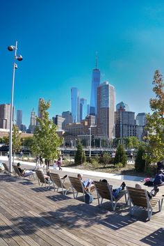 people are sitting on benches near the water in front of a cityscape with skyscrapers