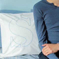 a person sitting on a bed holding a pillow with a dollar sign drawn on it