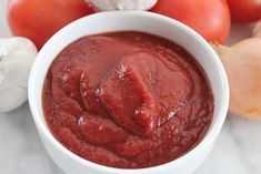 a white bowl filled with red sauce next to garlic and tomatoes