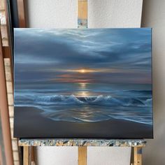 an easel with a painting on it and the sun setting in the sky over the ocean