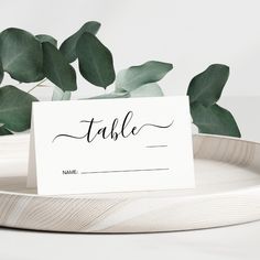 a place card sitting on top of a white plate next to a green leafy plant