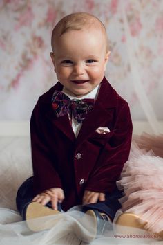 a smiling baby in a red suit and bow tie sitting on a white sheeted bed