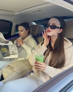 two women sitting in the back seat of a car, one holding a green drink